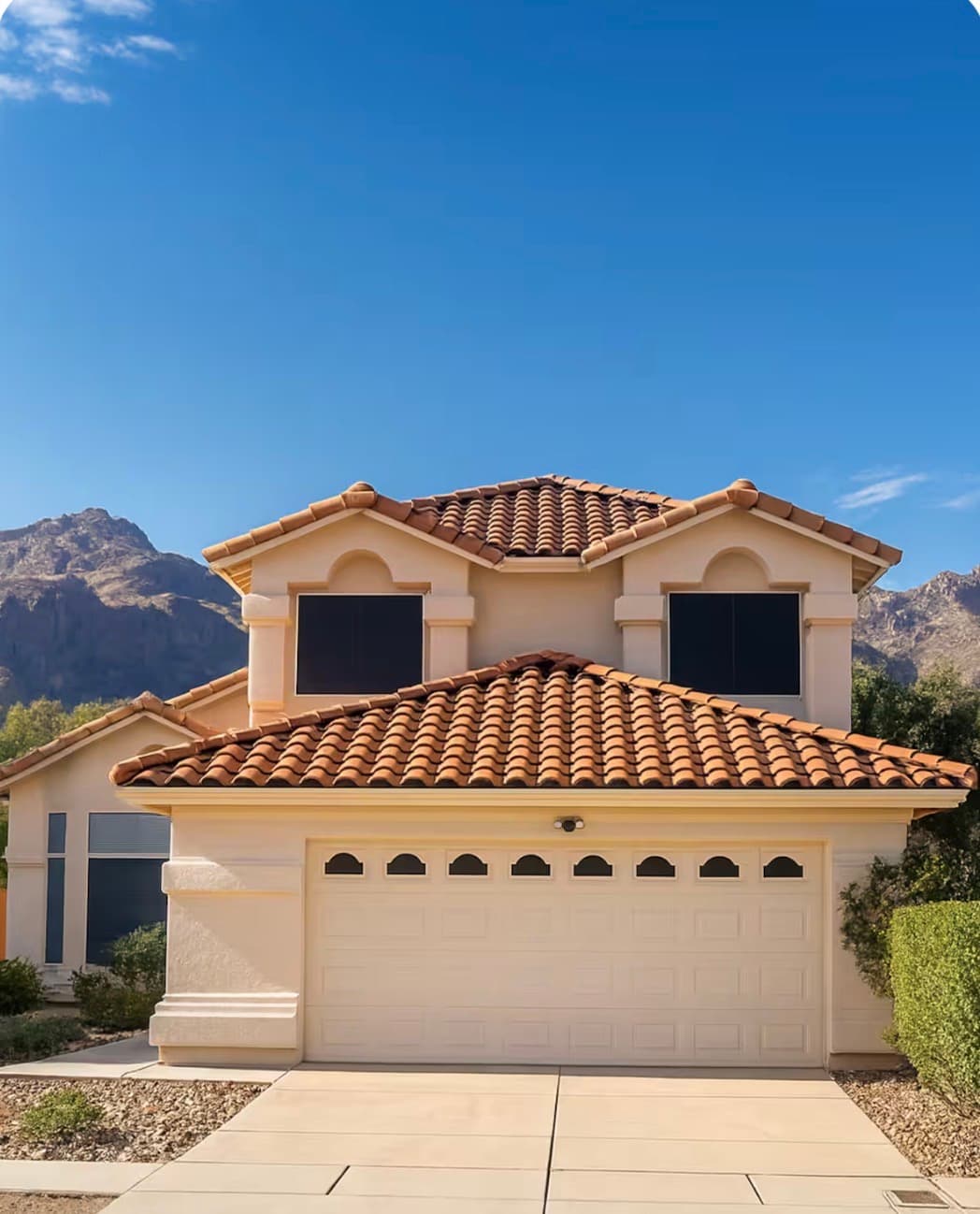 Tan stucco house with terracotta roof tiles and white garage door against desert mountain peaks.